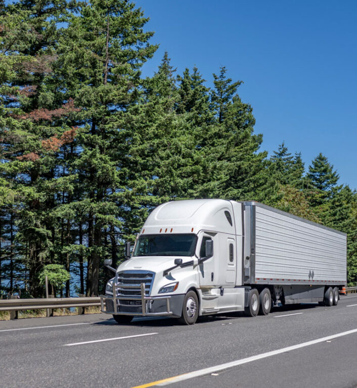 White semi truck driving down a forest covered highway transporting commercial cargo in reefer semi trailer.