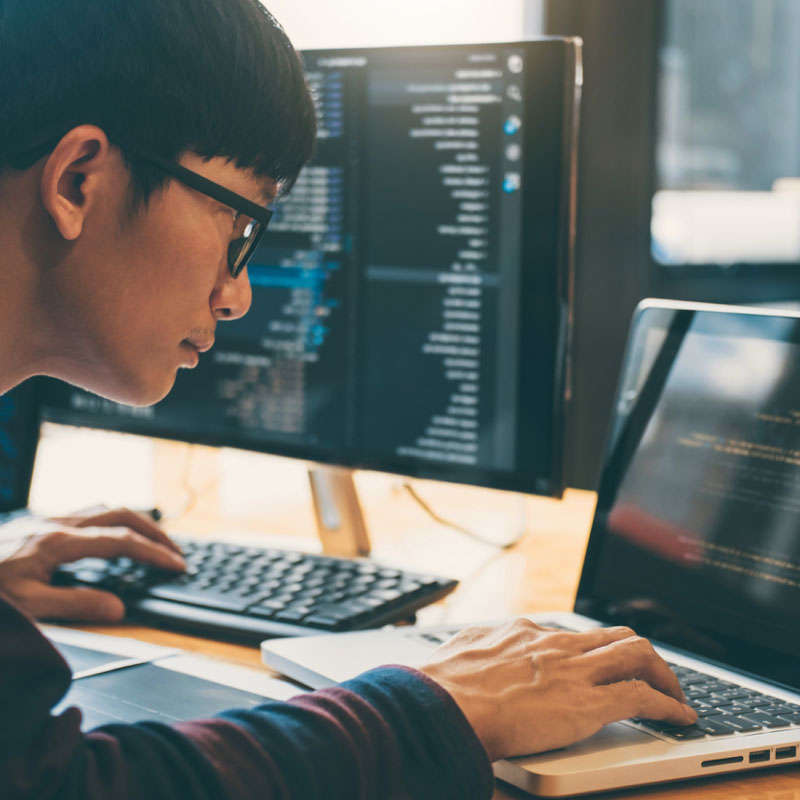 An engineer looking through code on both a laptop and desktop computer.