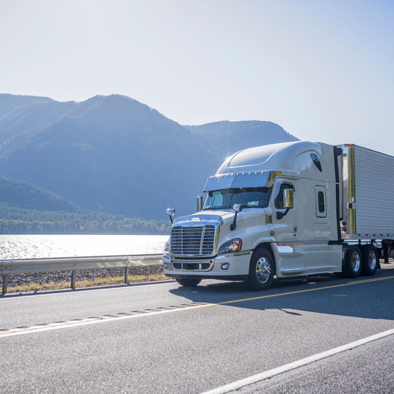 White semi truck driving down highway in front of mountains and water willed bay.