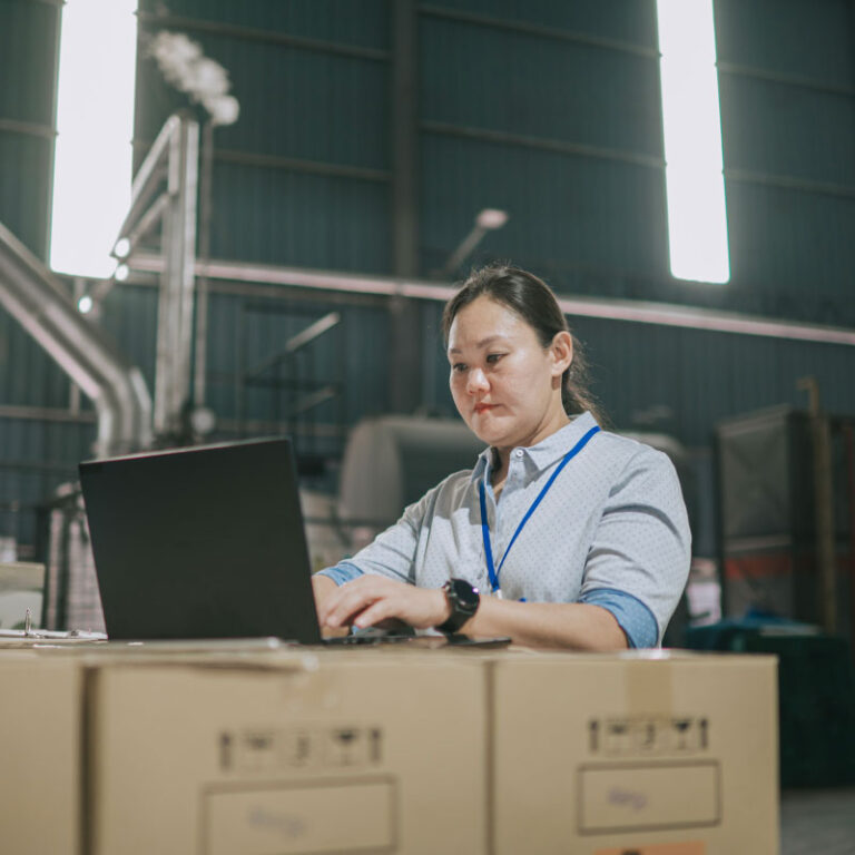 Woman working on laptop on pile of boxes in warehouse.