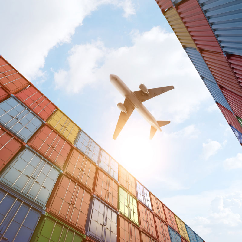 Cargo plane flying above a stack of shipping containers at a shipping port.