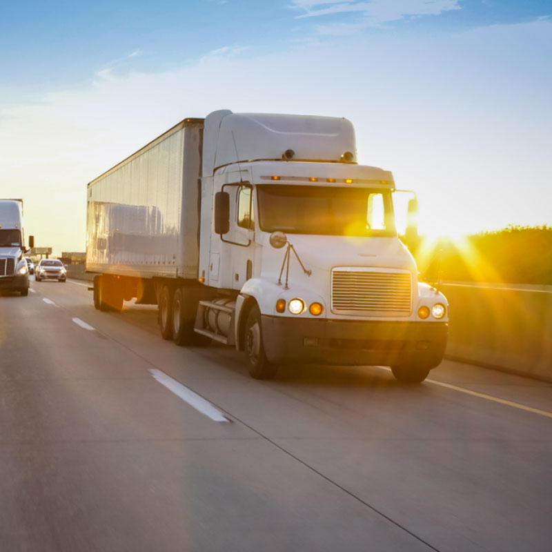 Two white semi trucks driving down the highway to their destination at sunset.