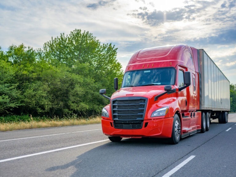 A classic big red semi truck transporting cargo on a road with a healthy green scenary.