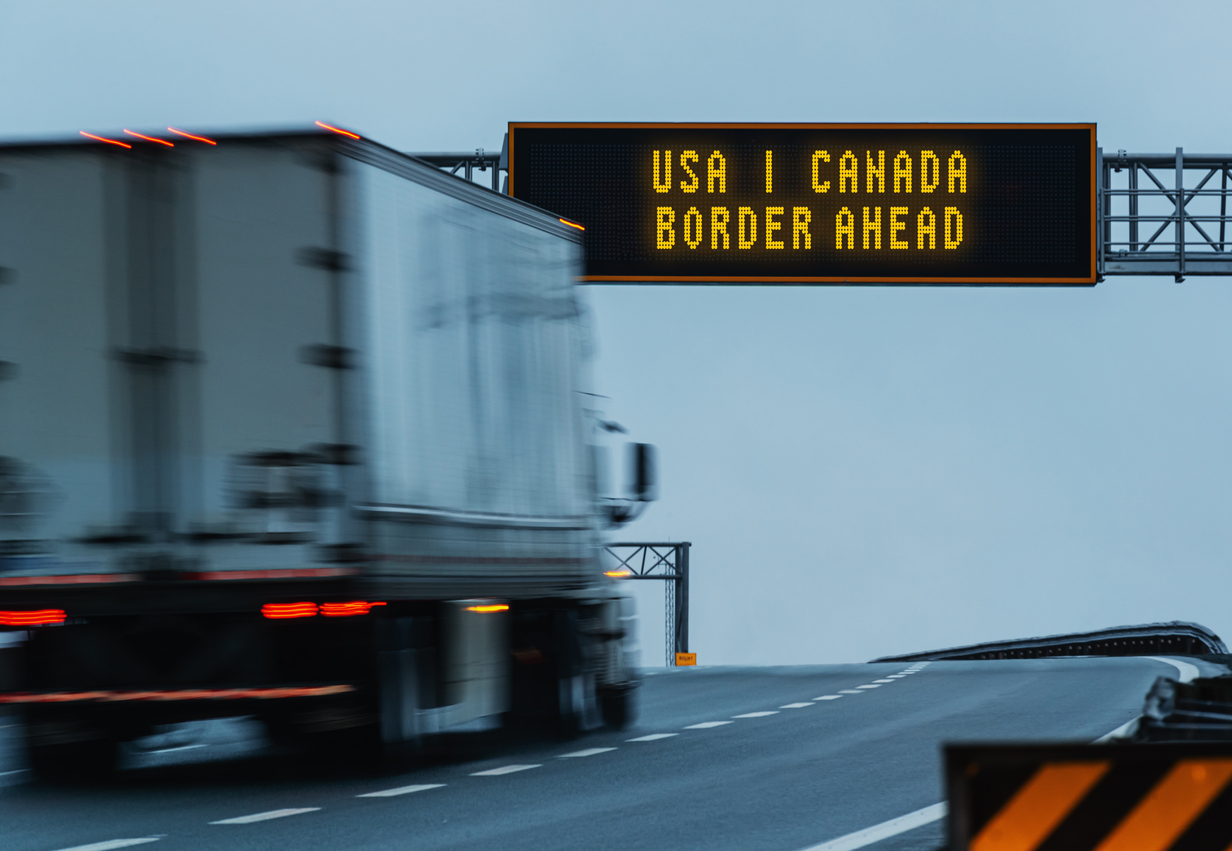 A semi truck driving under a highway sign that says "USA/Canada Border Ahead."